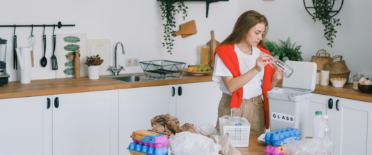 A woman in a kitchen sorting glass containers for recycling while surrounded by items to be organized, promoting sustainable practices.