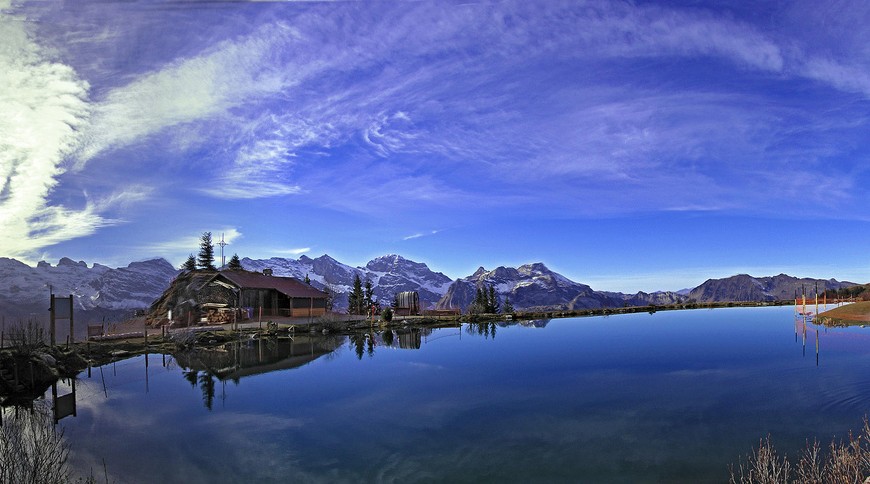 Il cielo si rifrette nel lago Härzlisee (Svizzera); sullo sfondo una baita e le catene montuose innevate