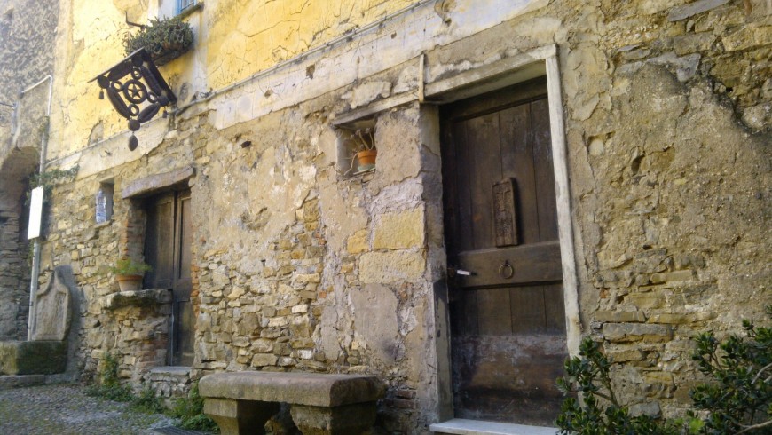 Edificio in pietra; la porta in legno reca un volto inciso e dal muro sporge (Colla Micheri, SV)