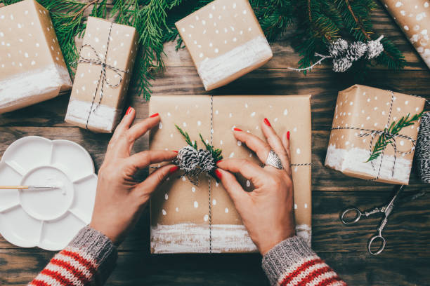 Le mani della donna confeziona i regali di Natale su carta marrone decorata con neve dipinta, rami di abete e pigne
