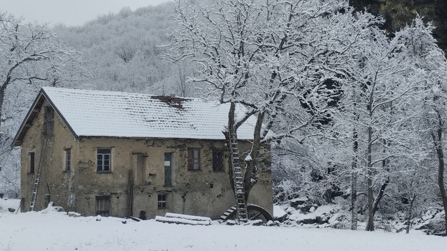 Antico mulino in pietra sotto la neve, circondato da alberi spogli e da un paesaggio invernale.