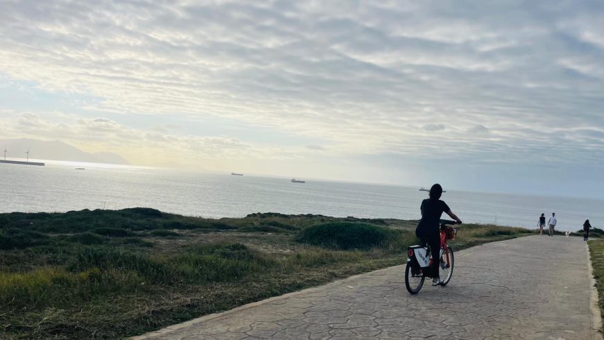 Costa di Bilbao con percorso ciclopedonale, persone in bicicletta e a piedi, mare sullo sfondo e pale eoliche all’orizzonte.