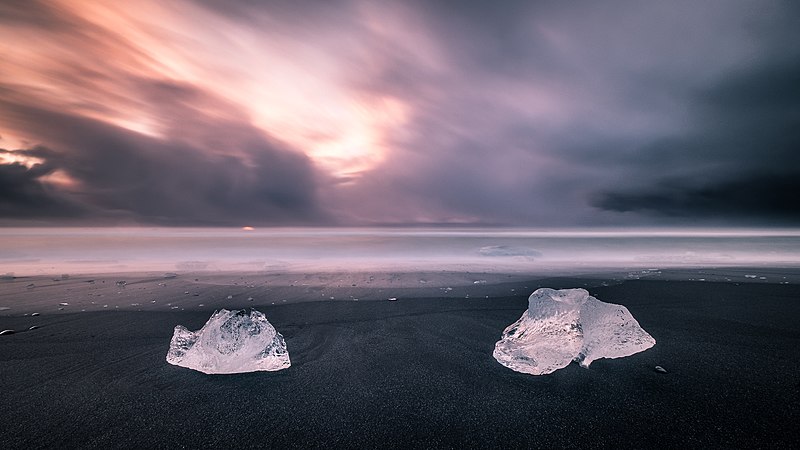 Blocks of ice on black sand, sea and pink sky in the background