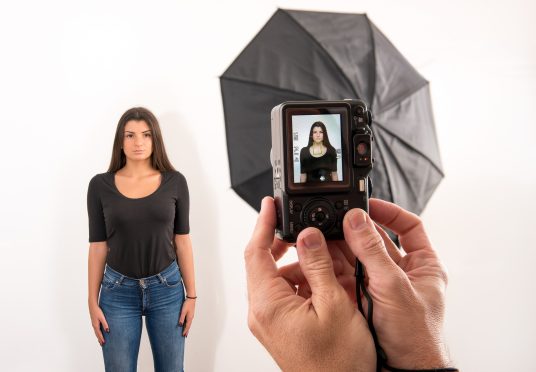 Attractive woman posing for her passport photo in a photographic studio with the photographers hands in the foreground holding the camera with viewfinder visible