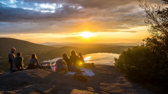 A small group of people sitting on top of a hill looking at a sunset in the distance.