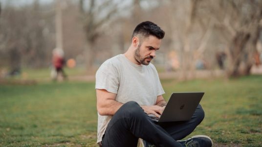 A man sitting on the grass in a park while using his laptop.