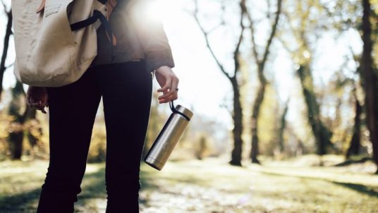 A person walking in the park carrying a backpack and a reusable thermos