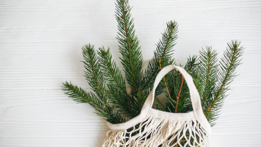 Net shopping bag with winter decorations, zero waste holidays. Sustainable lifestyle. Reusable cotton bag with green spruce branches on white rustic background.