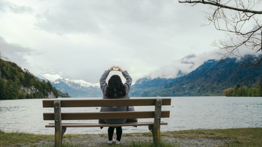 A woman on a bench by a lake forming a heart shape with hands above her head