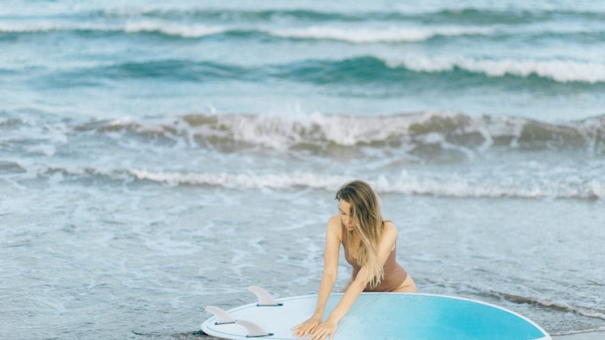 Woman in brown bathing suit, holding hands over blue surfboard, sea, ocean