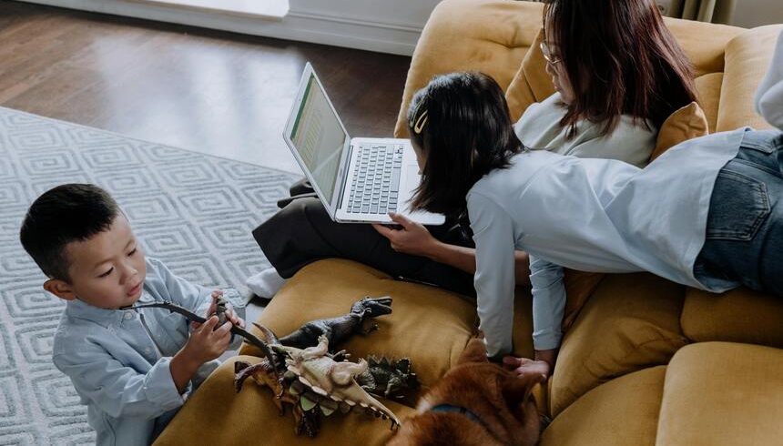 A parent using a laptop with children and a dog on a couch.