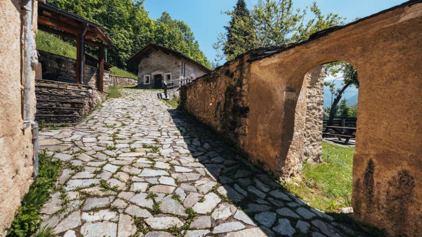 A sunlit stone path runs through a quiet alpine village, with a rustic wall and arch framing greenery and traditional houses.