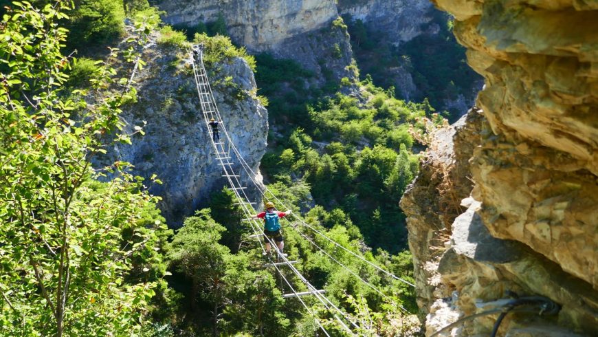 A narrow suspension bridge crosses a rocky gorge, surrounded by lush vegetation and dramatic cliffs.