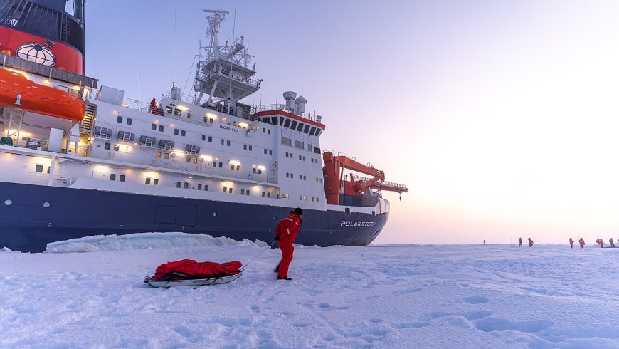 Scientist standing on Arctic sea ice in front of the research vessel RV Polarstern during a scientific expedition