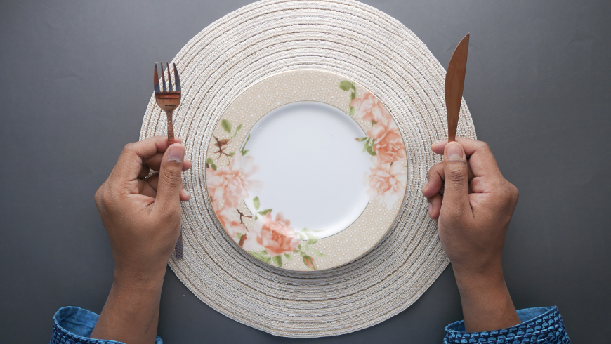 Person sitting at a table holding a fork and knife in front of an empty plate, symbolizing hunger and appetite control.