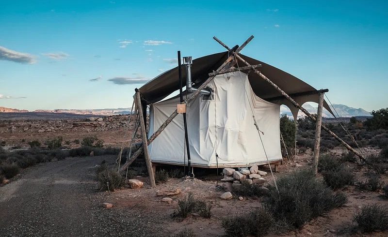 Ein luxuriöses Glamping-Zelt in einer kargen Wüstenlandschaft unter einem klaren Abendhimmel. Das Zelt ist mit einer robusten Plane und Holzpfosten konstruiert und verfügt über ein sichtbares Ofenrohr.