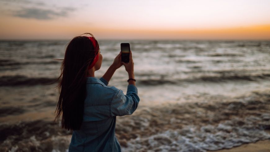 Junge Frau fotografiert das Meer bei Sonnenuntergang mit ihrem Smartphone, um am Ecobnb-Regenerate-Wettbewerb teilzunehmen.